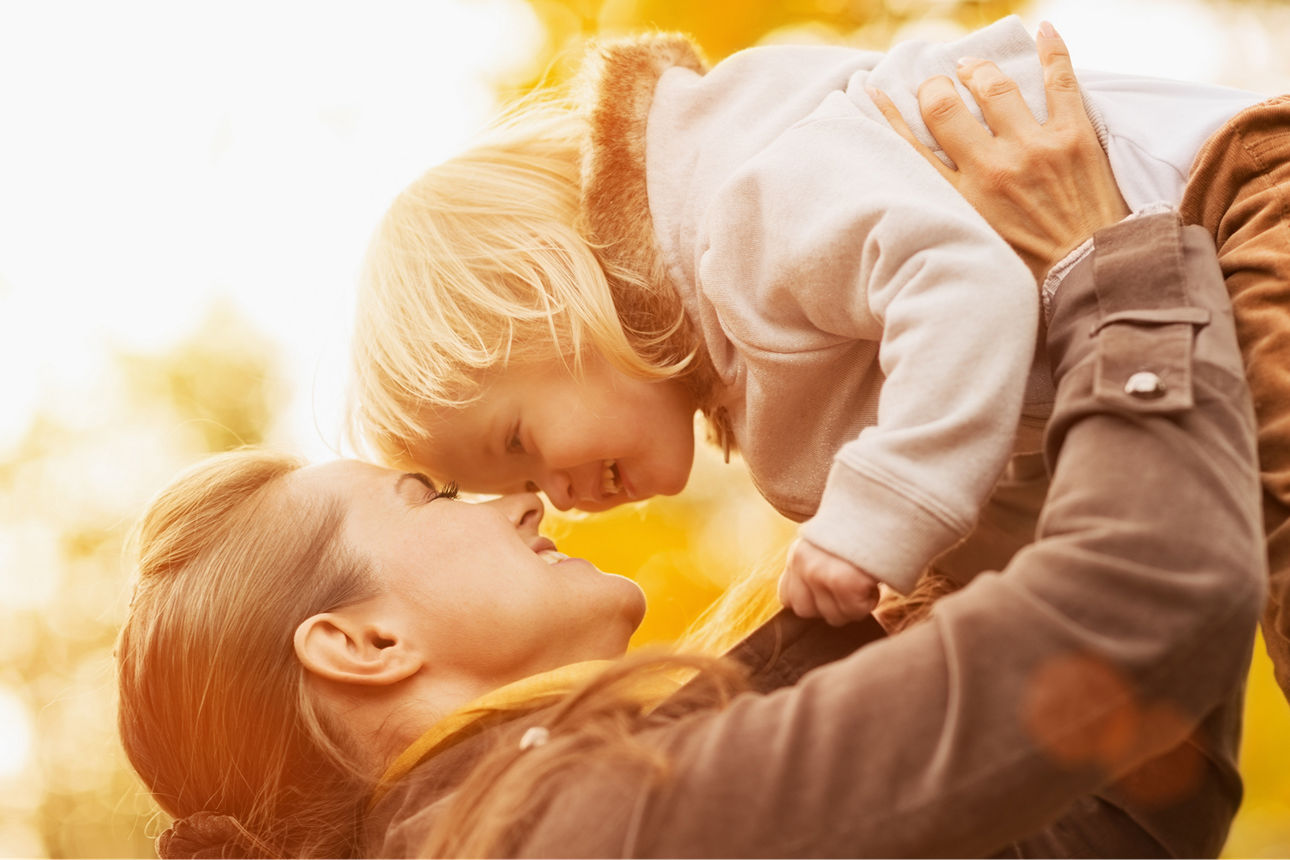 Mutter hält Kleinkind über sich in die Höhe, ihre Köpfe berühren sich und sie lächeln sich an