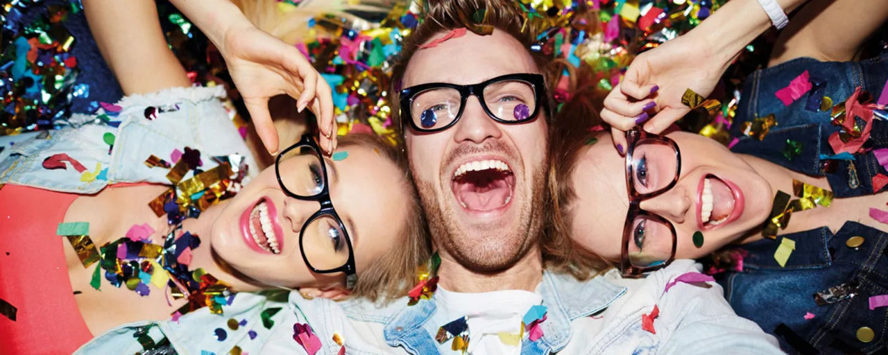 Drei Schulabgänger mit Brille sind bedeckt mit Konfetti und liegen lachend auf dem Rücken.
