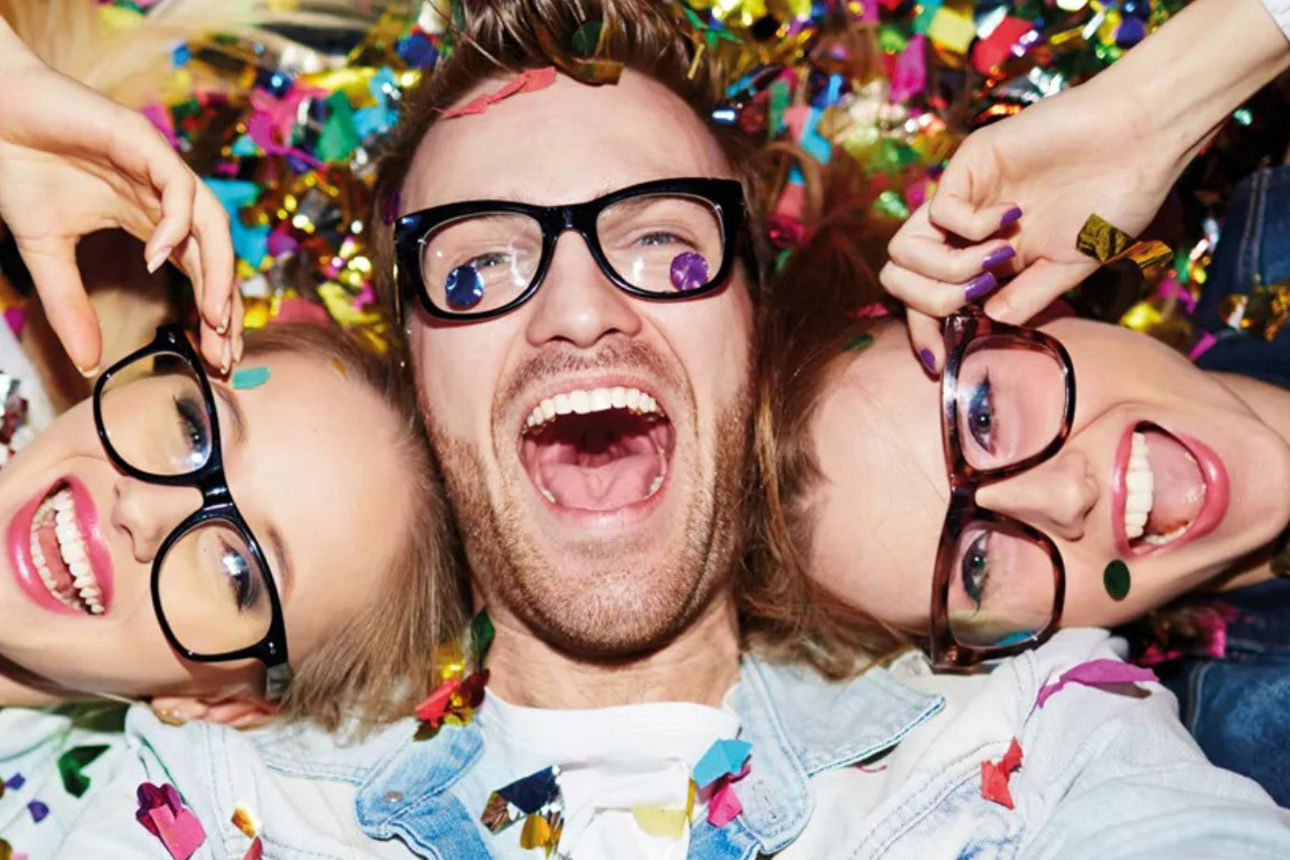 Drei Schulabgänger mit Brille sind bedeckt mit Konfetti und liegen lachend auf dem Rücken.