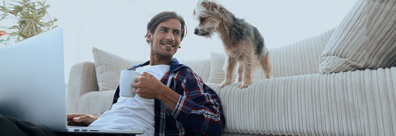 Junger Mann mit Kaffeetasse sitzt am Laptop und sein Hund schaut vom Sofa zu