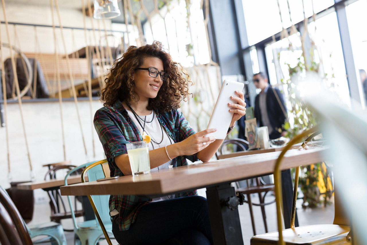 Eine junge Frau sitzt an einem Tisch in einem Cafe. Sie schaut auf ihr Tablet, dass sie in den Händen hält.
