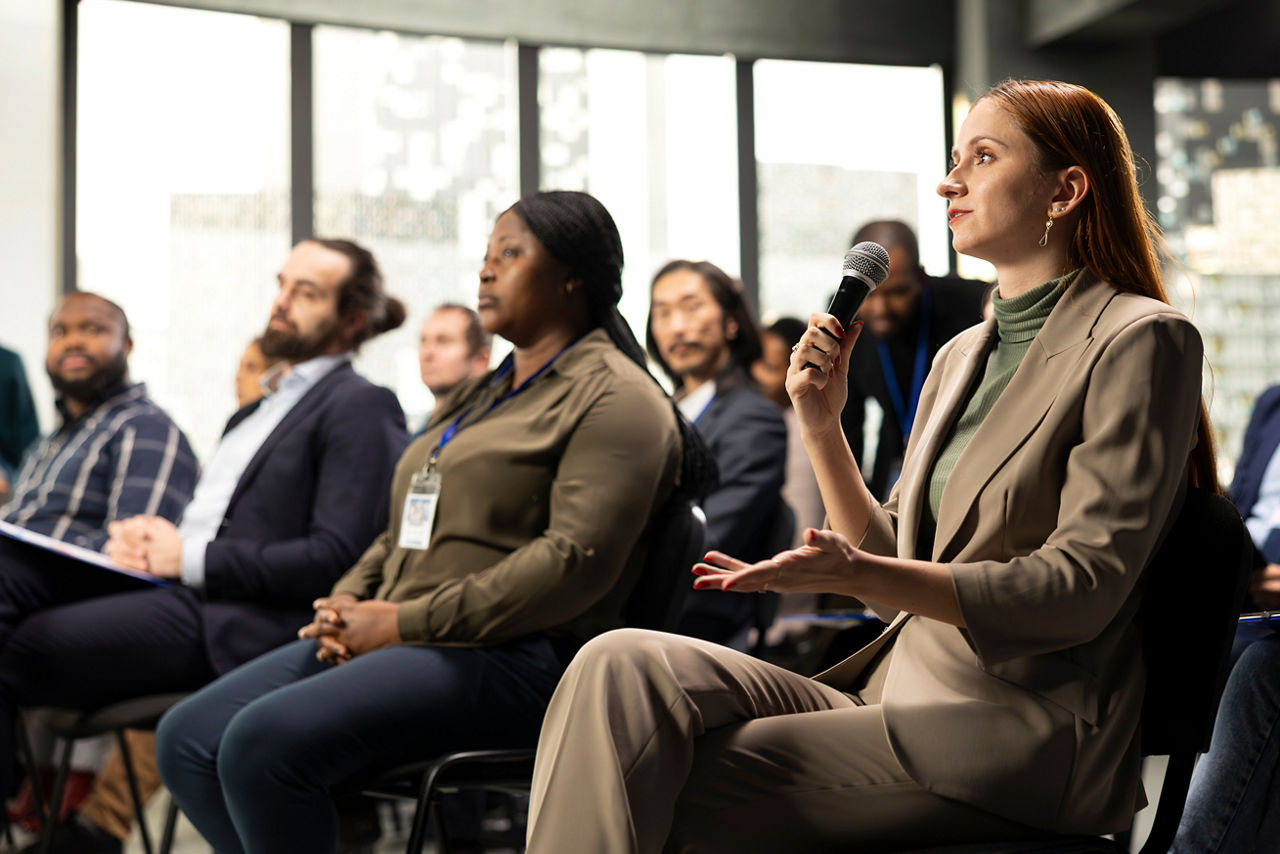 Young adult businesswoman in audience interacting with presenter, asking questions and engaging in debate. Mixed age participants include managers, employees and investors at summit.