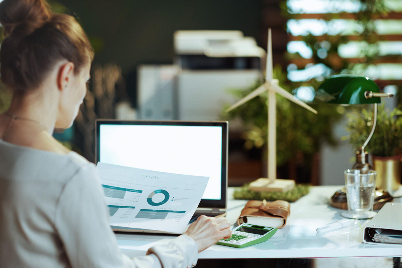 Frau am Laptop und Unterlagen am Arbeitstisch sitzend.