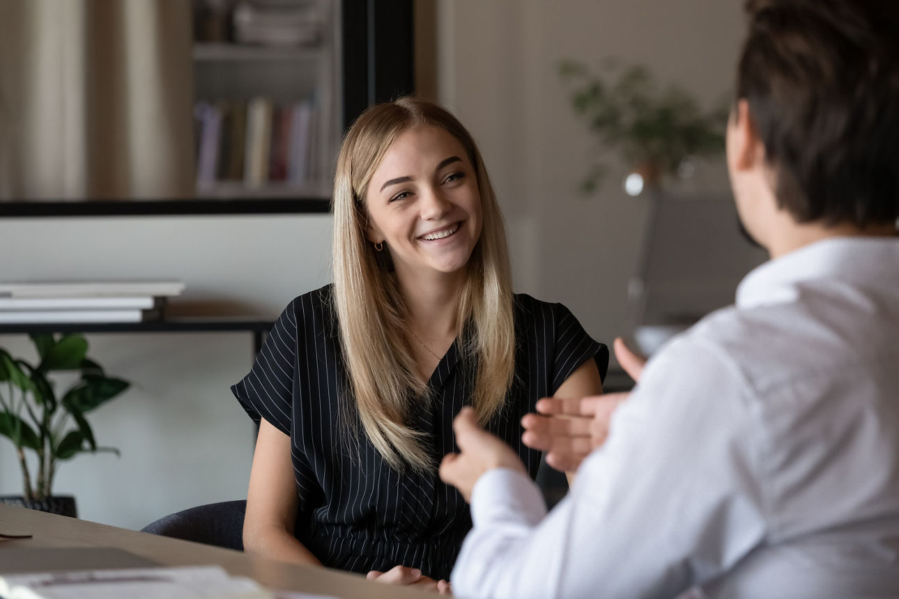 In der Mitte sitzt eine Frau seitlich an einem Tisch. Ihr gegenüber, mit dem Rücken zum Bild, sitz ein Mann. Die beiden unterhalten sich in einem Büro. Er gestikuliert, sie lächelt.