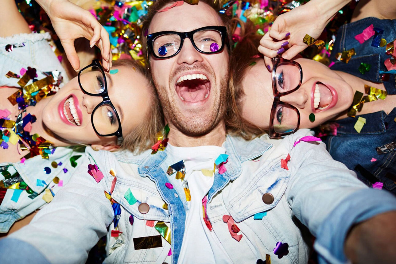 Ein lachender Mann, an dessen Kopf je Seite eine lachende Frau liegt. Die drei Brillenträger sind mit Konfetti überschüttet.