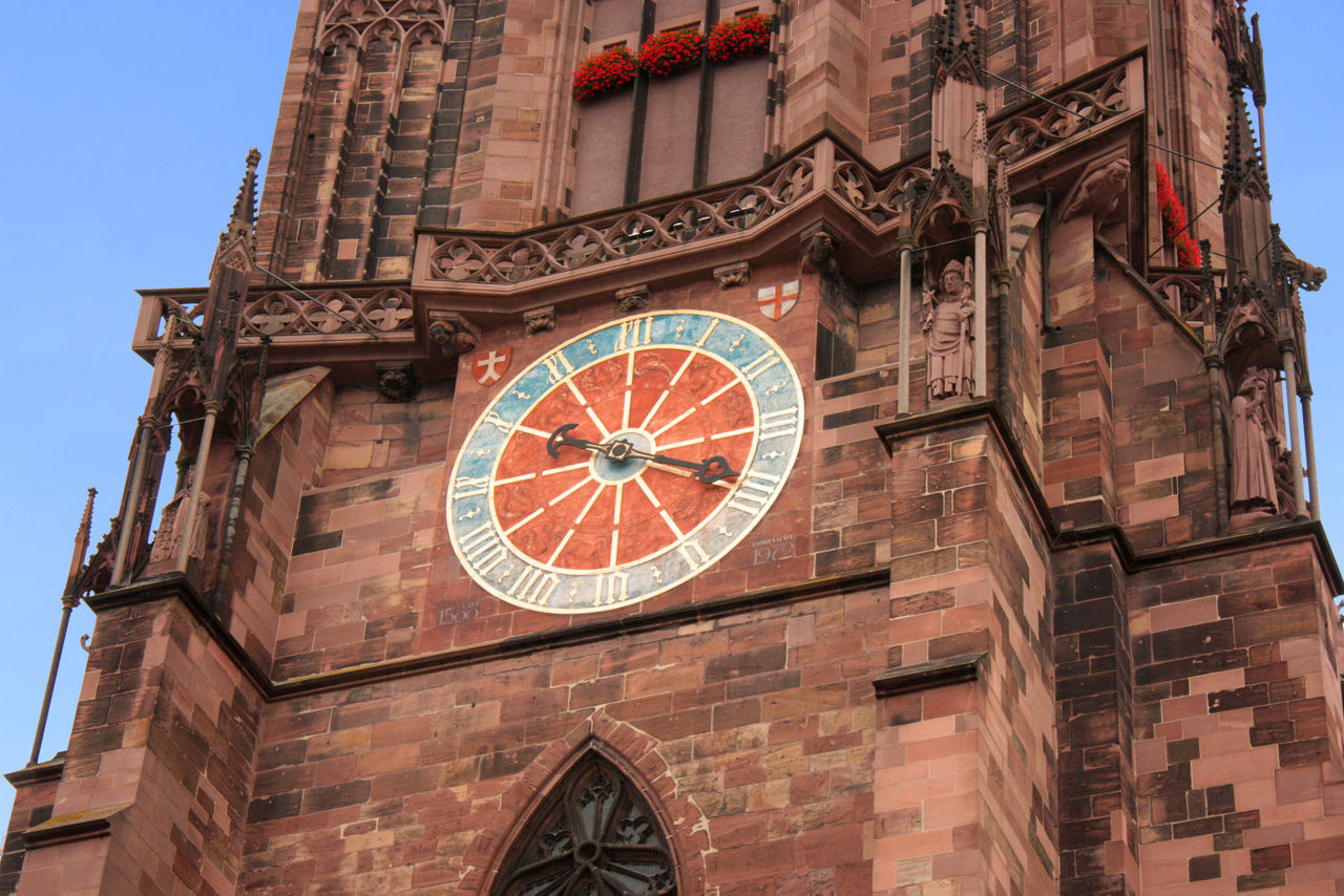 Eine bunte Uhr mit römischen Ziffern ist an einem roten Sandsteinturm mit gotischen Verzierungen angebracht.