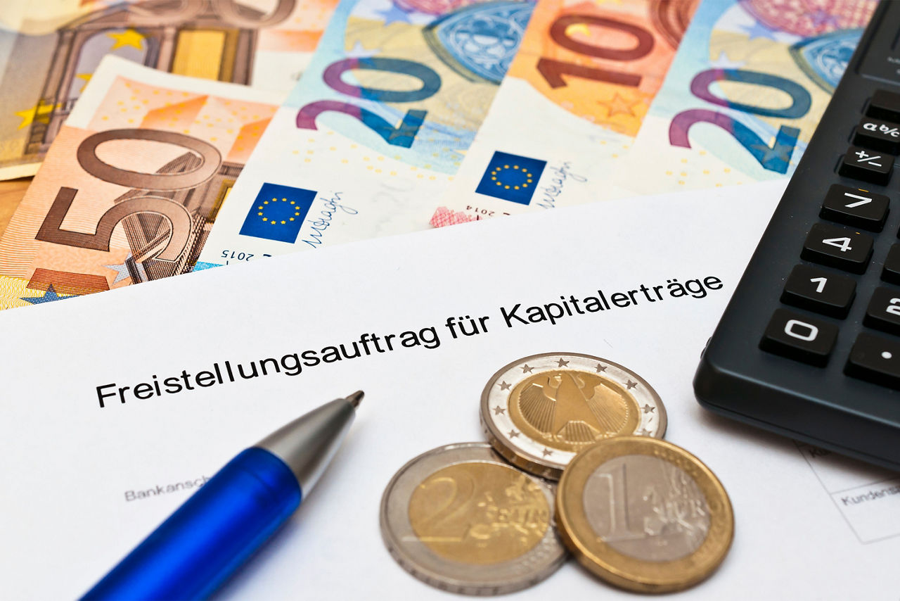 Das Bild zeigt ein Blatt Papier mit der Aufschrift "Freistellungsauftrag für Kapitalerträge" das auf einigen Euro-Scheinen liegt. Auf dem Blatt liegen ein paar Euro-Münzen, ein Kugelschreiber und ein Taschenrechenr.