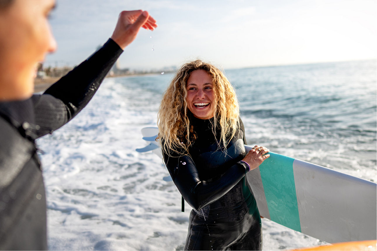 Person im Neoprenanzug steht mit Surfbrett im seichten Wasser, während der Freund daneben im Meer steht