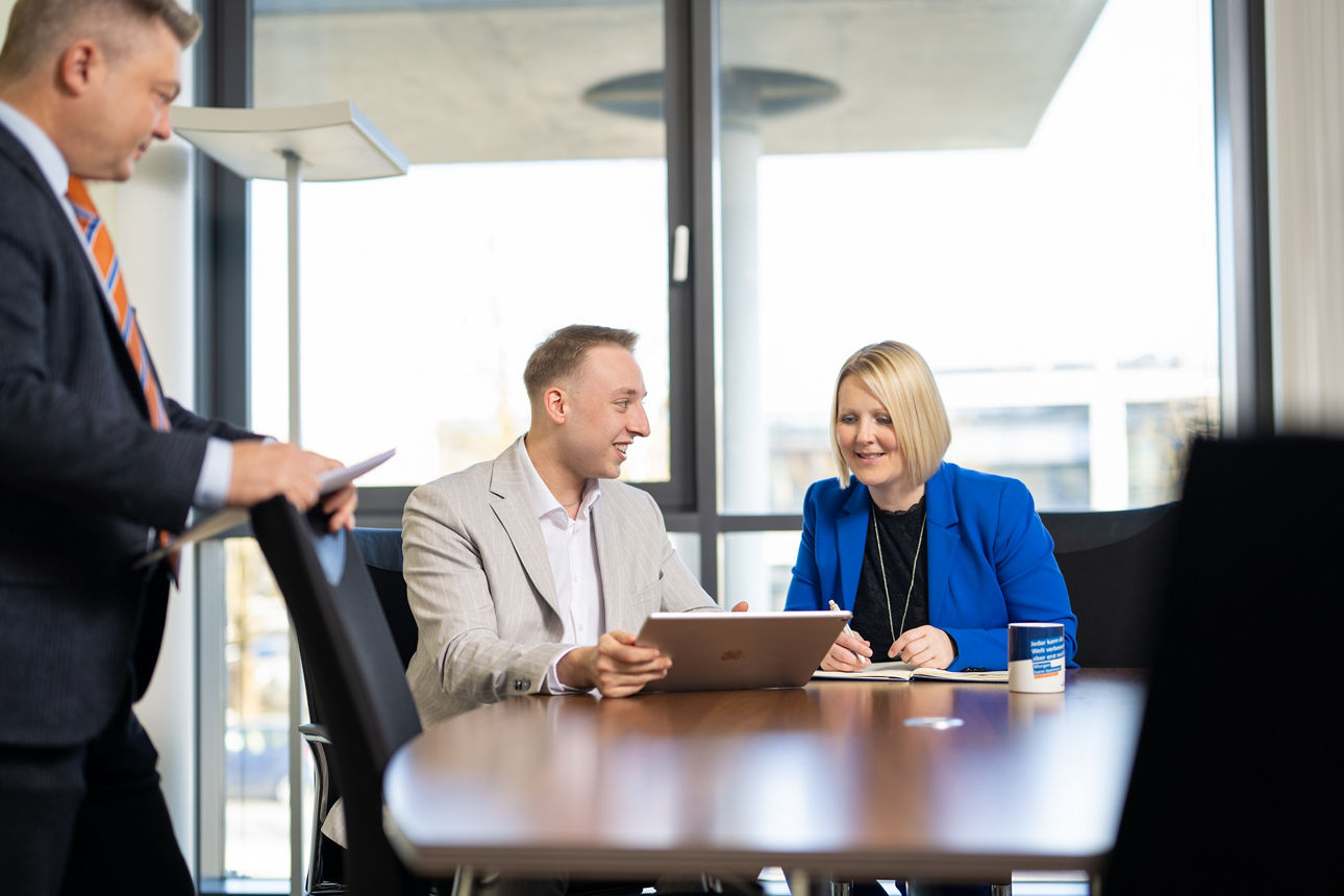 Drei Kollegen, ein Herr steht im Vordergrund links, ein Herr und eine Dame sitzend an Beratungstisch in einem Meeting und besprechen sich unter Zuhilfenahme eines Tablets in modernem hellem Büro