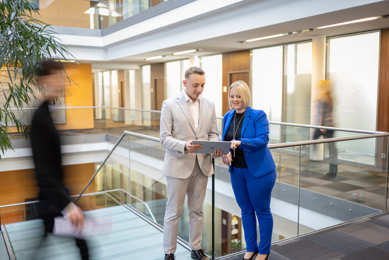 Junger Mann in hellem Anzug und junge Frau in blauem Hosenanzug stehen in modernem Bankgebäude und blicken auf ein Tablet. Im Hintergrund verschwommen die helle Innenarchitektur der Bank