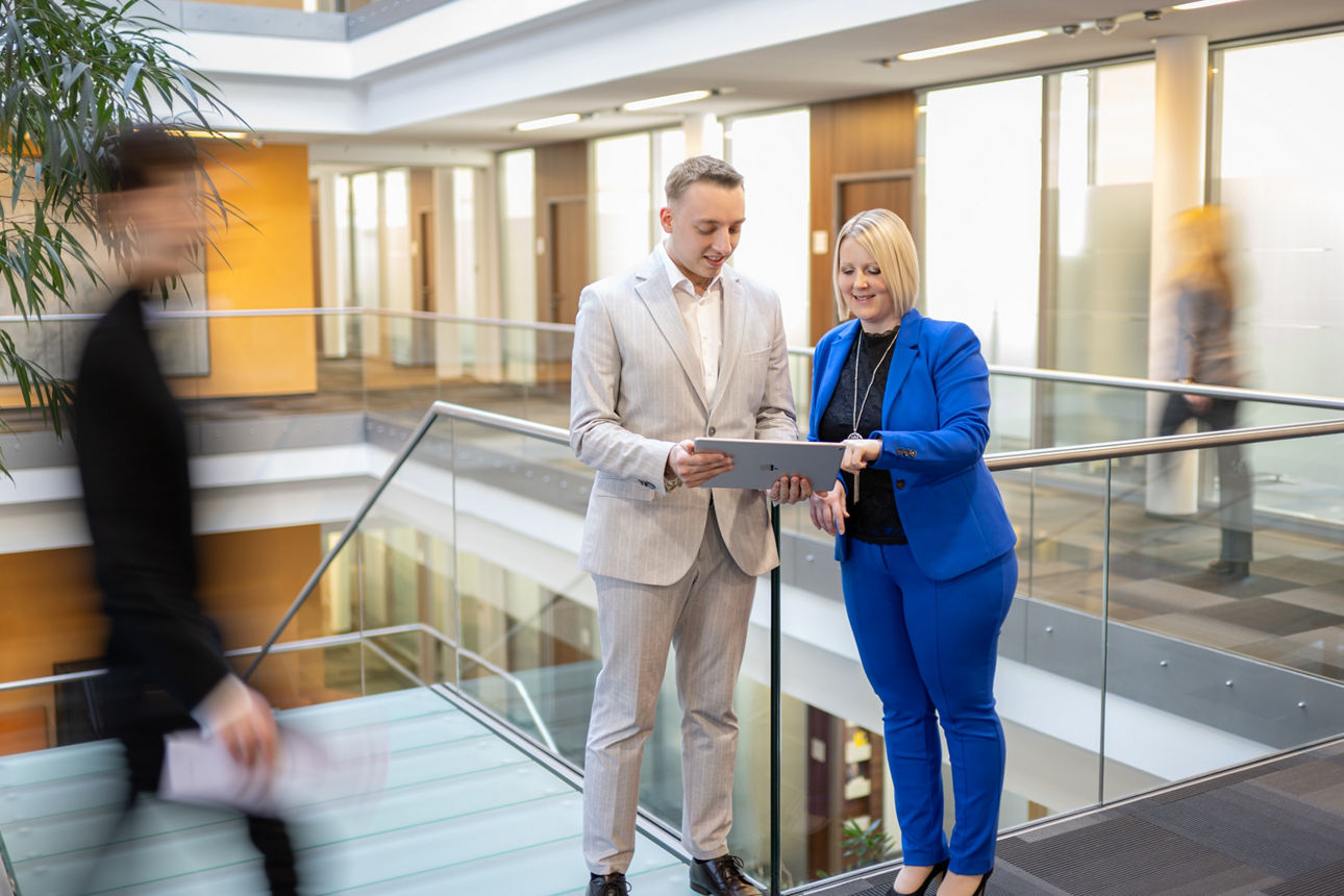 Junger Mann in hellem Anzug und junge Frau in blauem Hosenanzug stehen in modernem Bankgebäude und blicken auf ein Tablet. Im Hintergrund verschwommen die helle Innenarchitektur der Bank