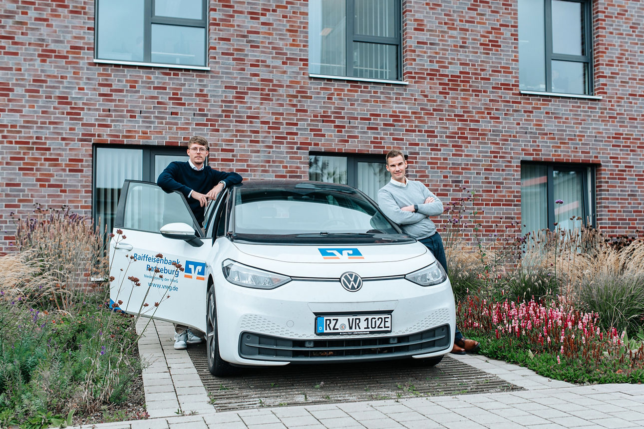 Zwei Männer lehnen sich lässig - jeweils rechts und links - an einem VReG E-Auto an.