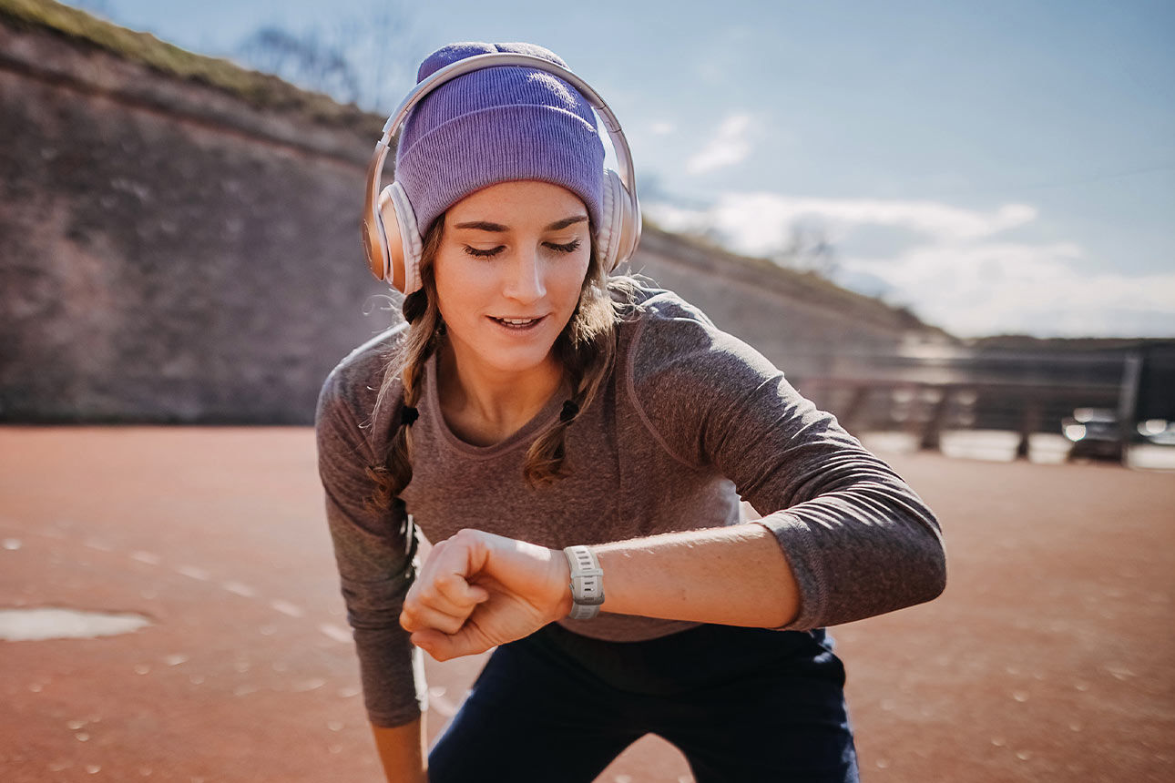 Junge Frau mit Kopfhörern schaut auf die Knie abgestützt nach einem Lauf auf Ihre Garmin Smartwatch