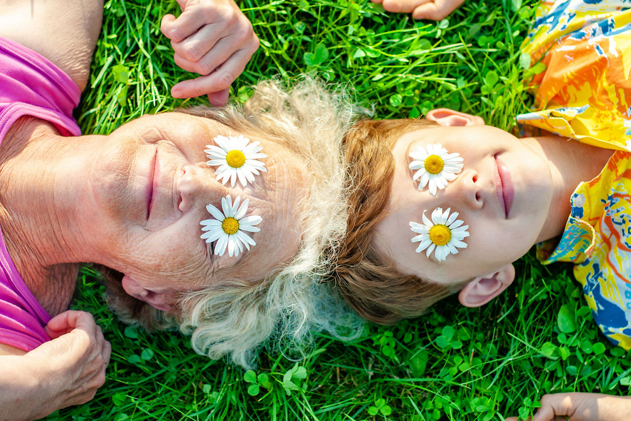 Ältere Dame und Kind liegen mit Löwenzahn bedeckt auf den Augen, auf einer Wiese