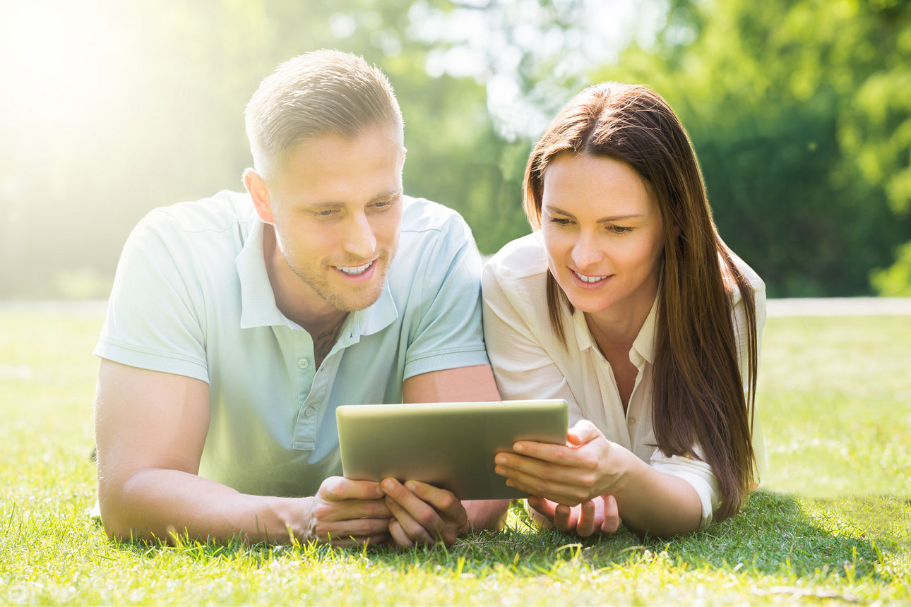 Junges Paar liegt im Gras und nutzt ein Tablet im Park.