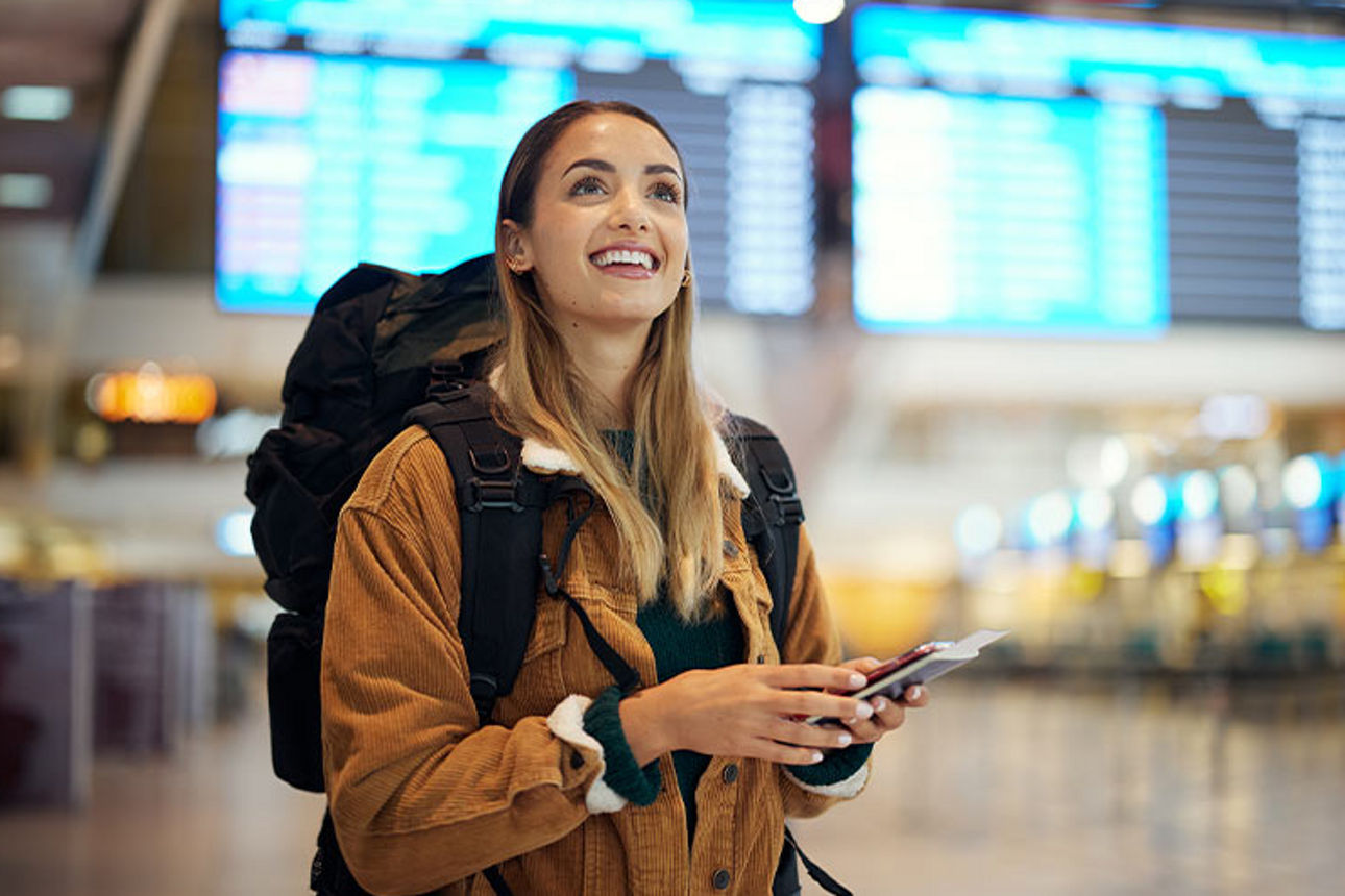 Junge Frau steht mit Gepäck und Reisepass in der Hand in der Schalterhalle eines Flughafens