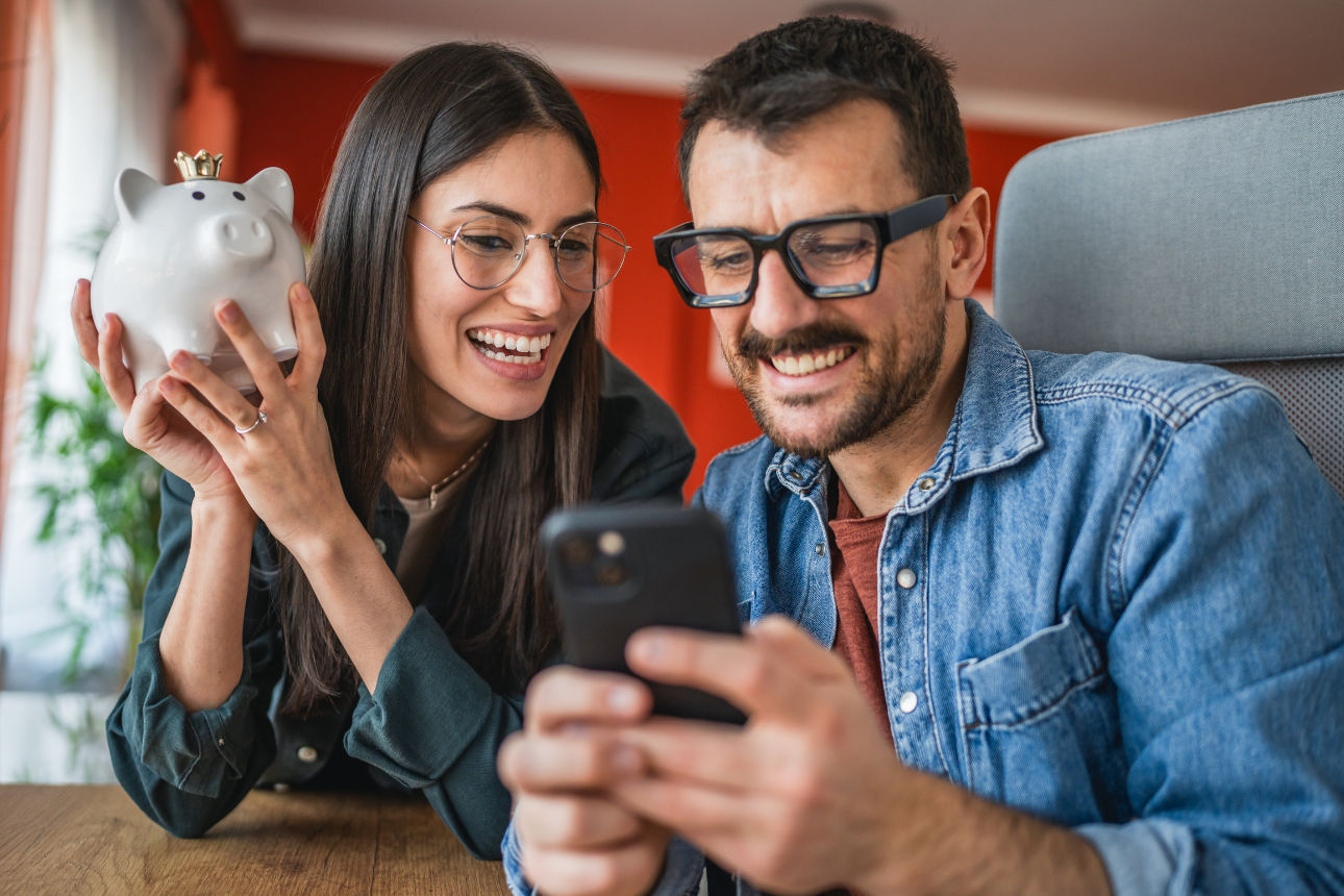Zwei Personen schauen gemeinsam auf ein Smartphone; eine hält ein Sparschwein, im Hintergrund Innenraum.