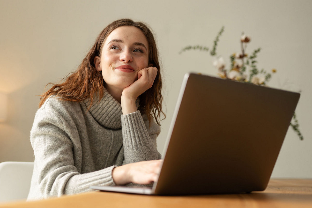 Junge Frau sitzt verträumt am Schreibtisch vor ihrem Laptop