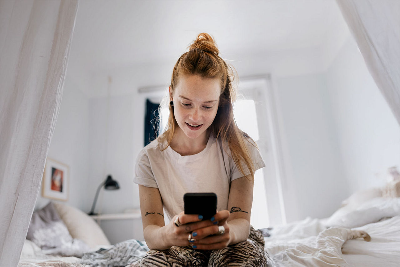 Junge Frau sitzt an der Bettkante und schaut gespannt in ihr Handy mit einem Hinweis "Garantierter Gewinn ab 10 Losen"