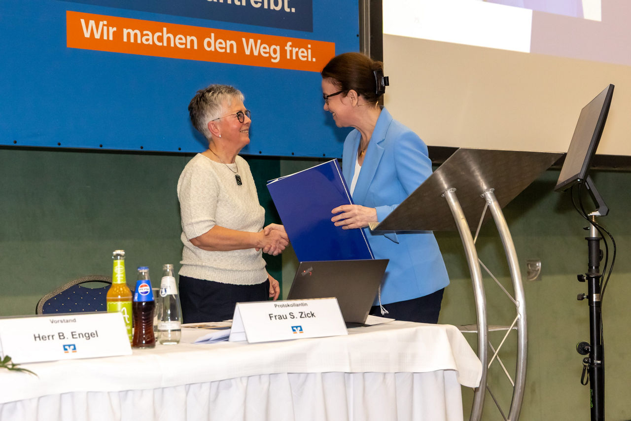  Menschen an einem Tisch, es wird eine Urkunde übergeben, im Hintergrund ist eine Leinwand zu sehen. 