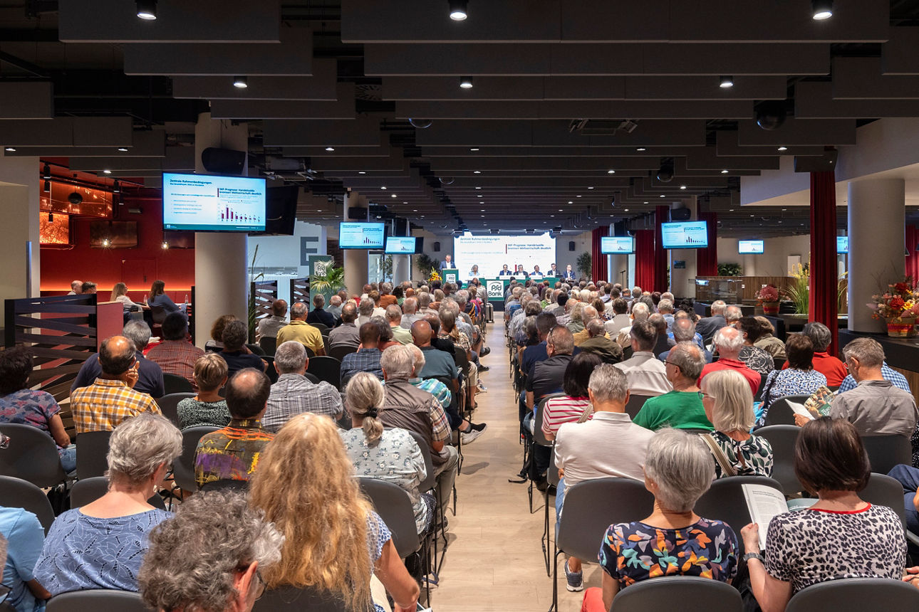 In einem Raum sitzen links und rechts Menschen in Sitzreihen und blicken nach vorne zur Bühne. Dort sitzen sechs Personen nebeneinander am Tisch, während eine weitere Person am Rednerpult steht. Im Hintergrund erkennt man eine Präsentation. 