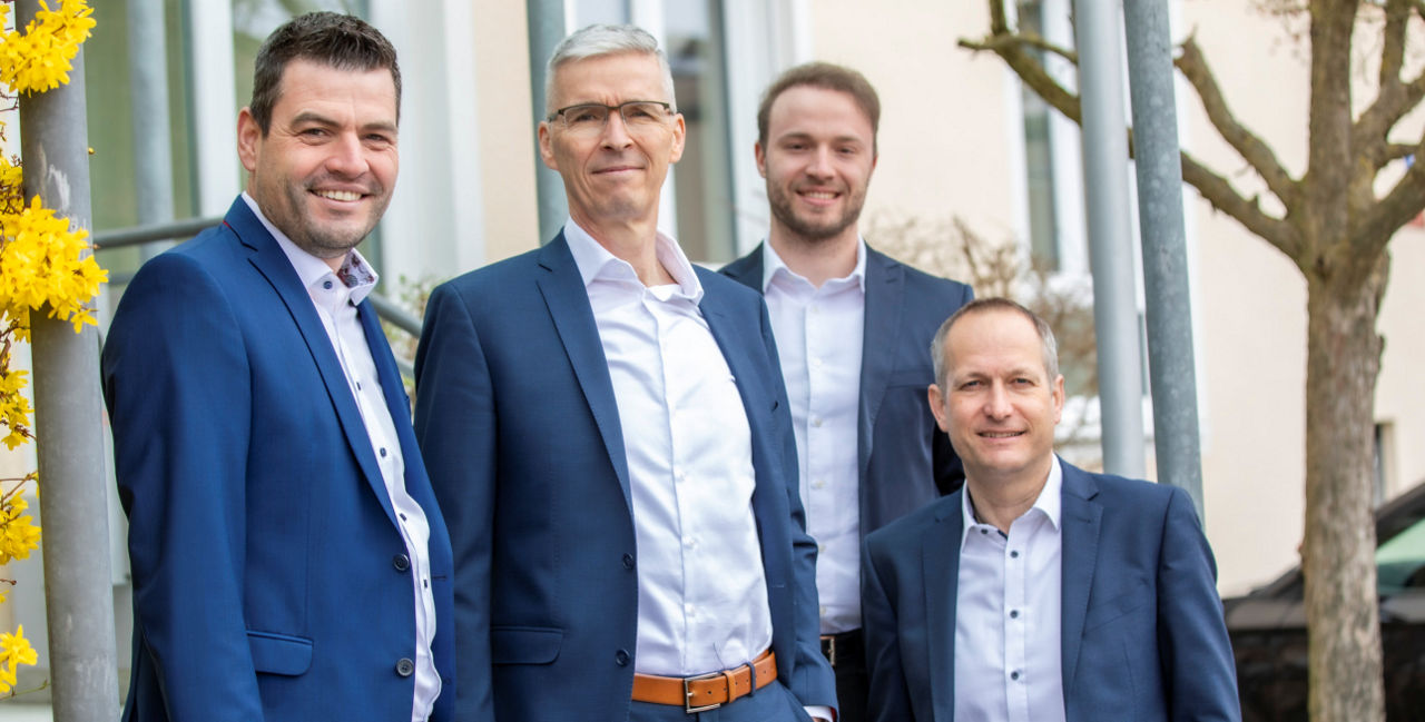 Vier Männer vor einem Haus zwischen zwei Bäumen