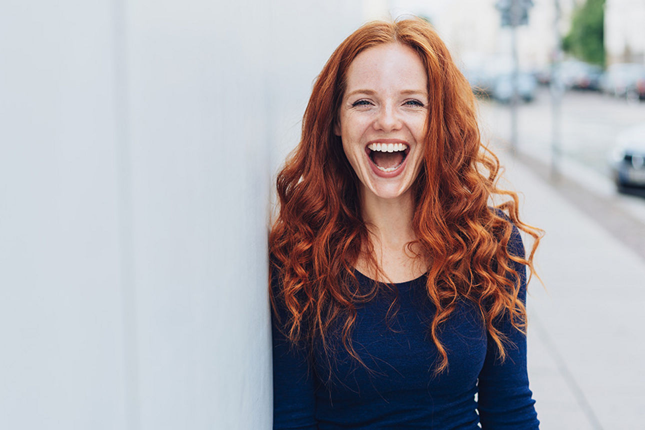 Rothaarige Frau lehnt gut gelaunt an einer Hauswand