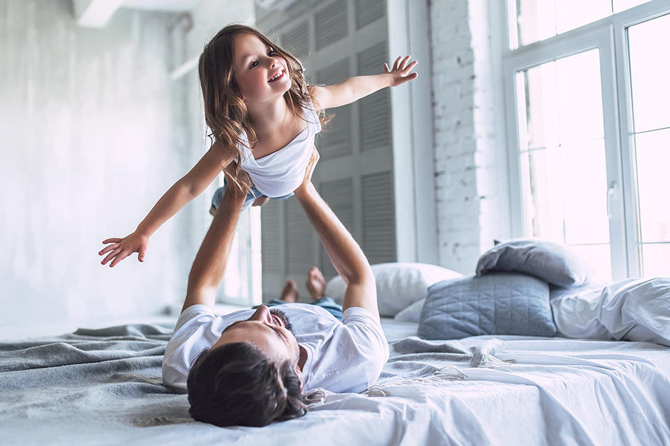 Vater liegt auf dem Bett und hält seine Tochter in die Luft, sie hat die Arme wie Flügel ausgebreitet