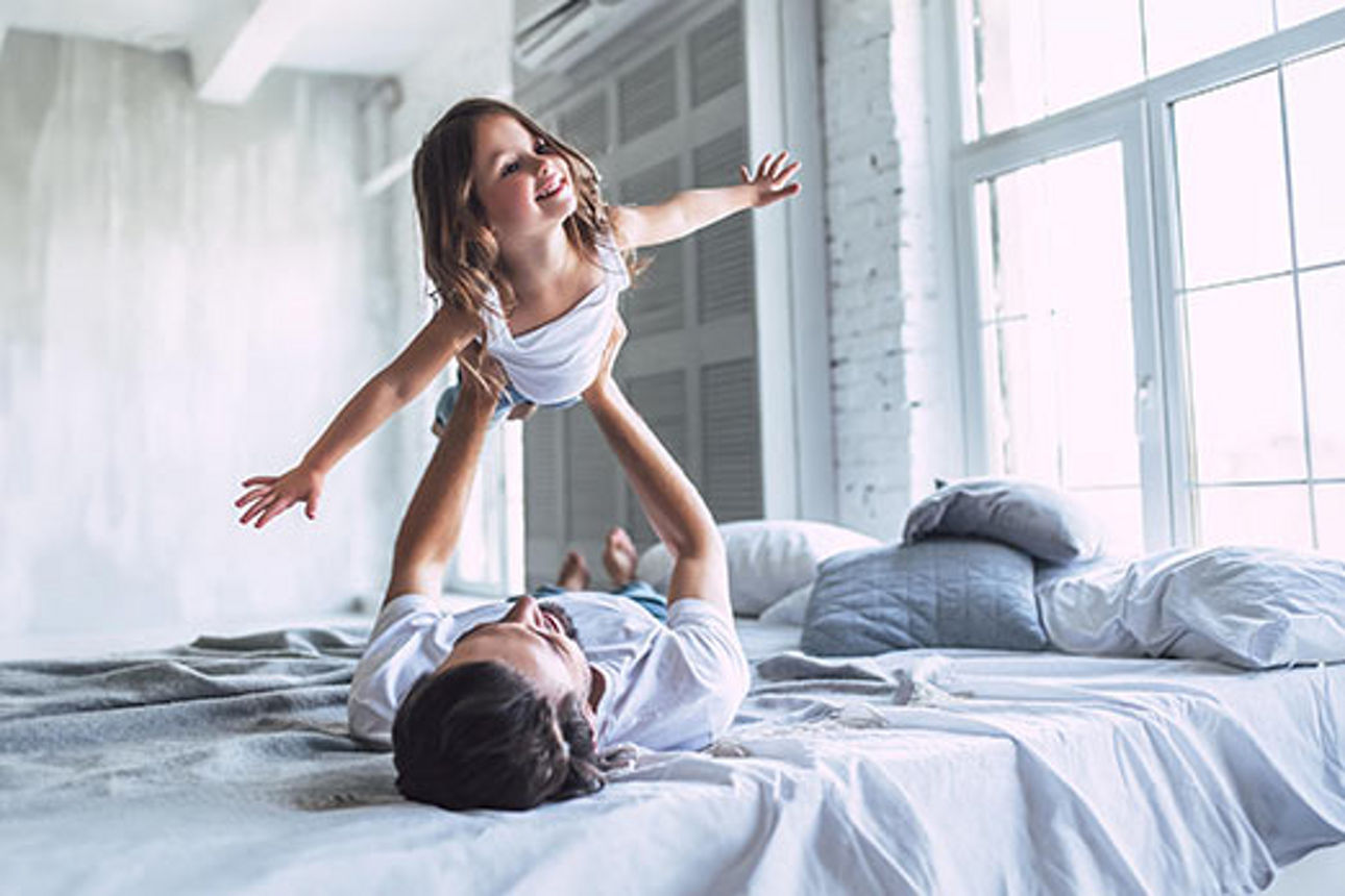 Vater liegt auf dem Bett und hält seine Tochter in die Luft, sie hat die Arme wie Flügel ausgebreitet