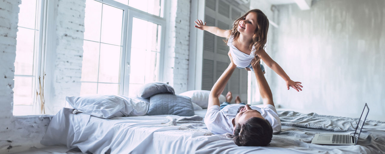Vater hält seine Tochter auf dem Bett liegend in die Luft, sie hat die Arme wie Flügel ausgebreitet