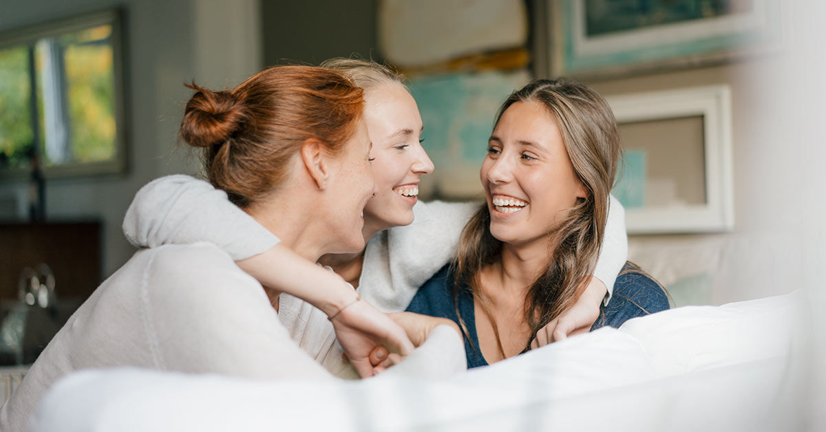 Drei junge Frauen sitzen lachend zusammen auf einem Sofa und genießen die gemeinsame Zeit.