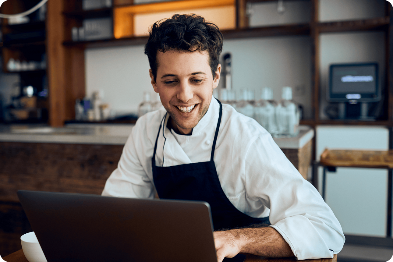 Ein junger Mann vor einem Laptop. Im Hintergrund ein Regal mit Flaschen