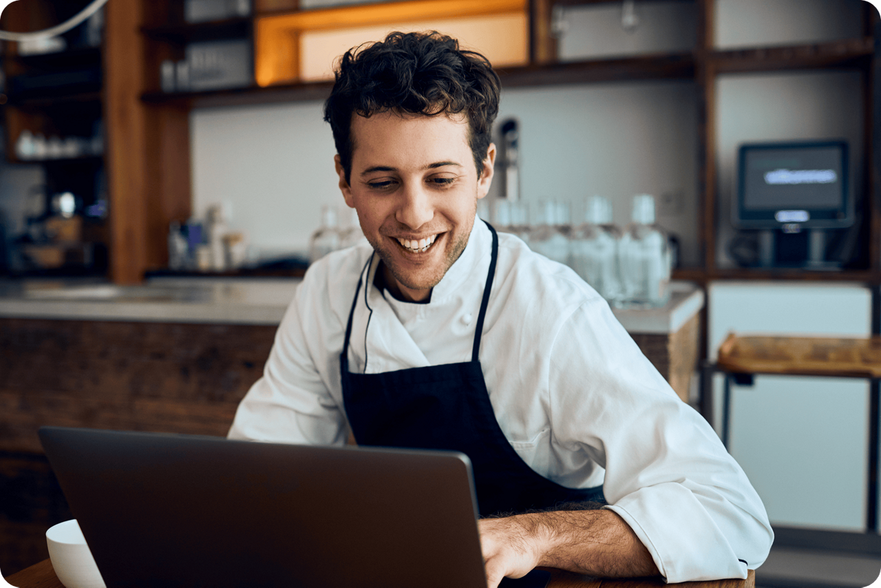 Ein junger Mann vor einem Laptop. Im Hintergrund ein Regal mit Flaschen