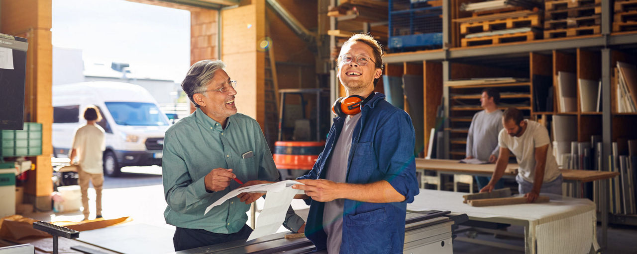 Älterer und jüngerer Mann stehen in einer Arbeitshalle und besprechen Arbeitsdokumente