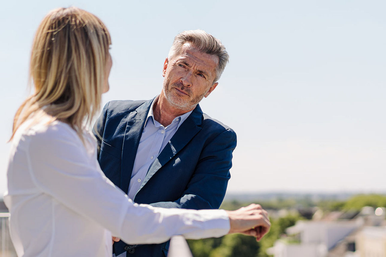 Ein Mann und eine Frau stehen am Geländer einer Dachterrasse und sehen sich an, im Hintergrund sind weitere Häuser erkennbar
