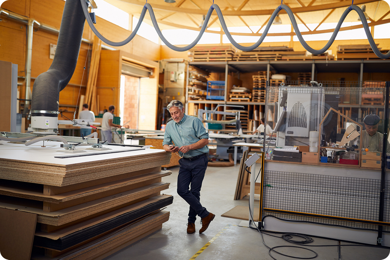 Mann steht in einer Arbeitshalle, lehnt lässig auf einem Stapel Spanplaaten und schaut auf sein Smartphone