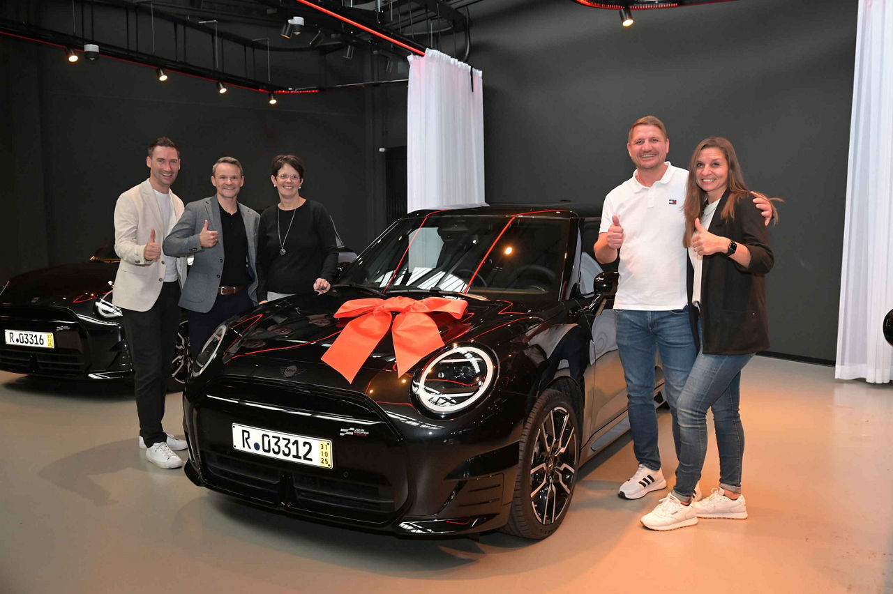 Das Bild zeigt fünf Personen, die in einem modernen Ausstellungs- oder Übergaberaum neben einem schwarzen Auto stehen. Auf de