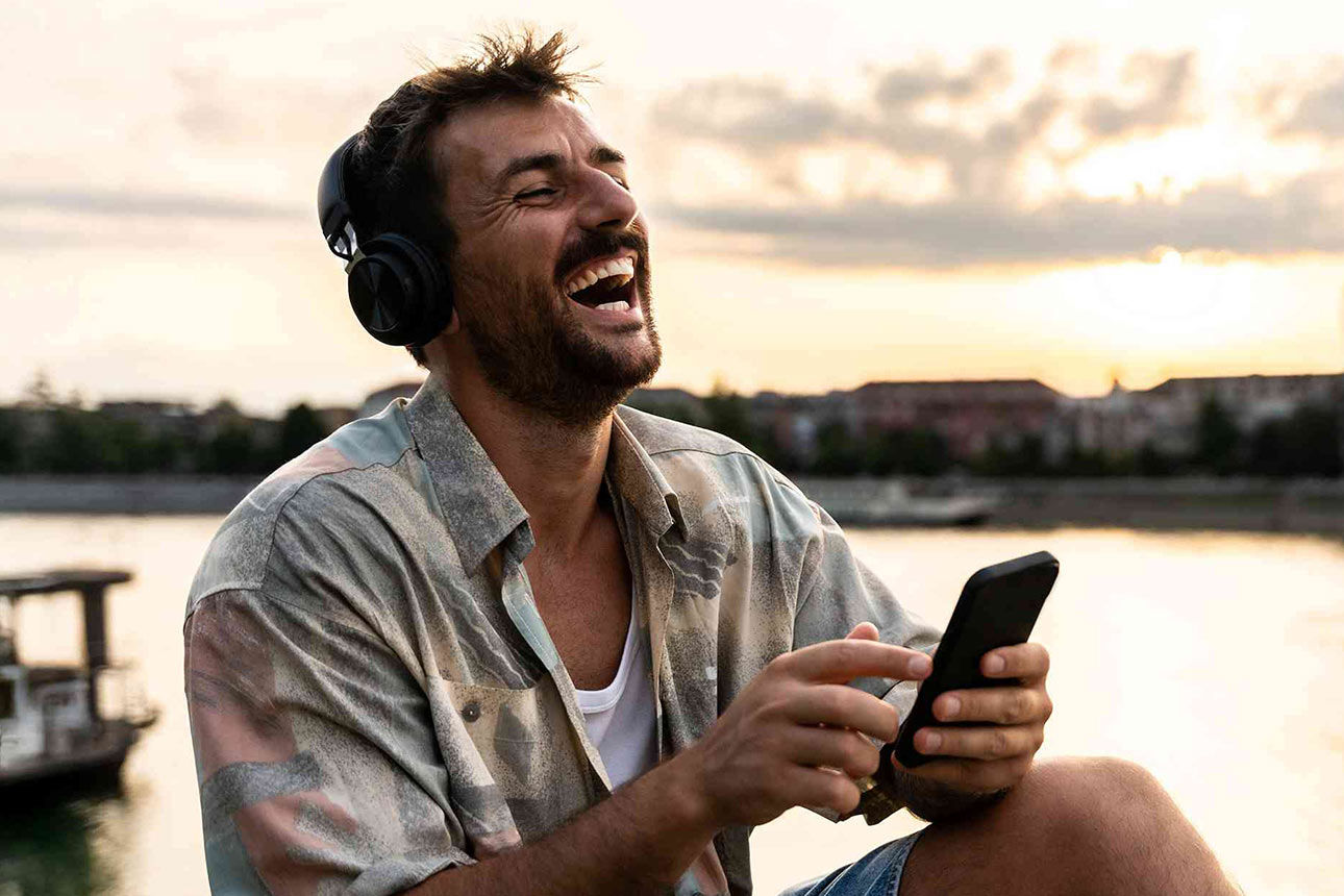 Mann mit Kopfhörer und Smartphone sitzt lachend am Flussufer.