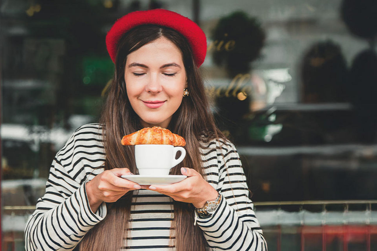 Eine Frau mit Baskenmütze hält eine Tasse Kaffee mit Croissant in den Händen.