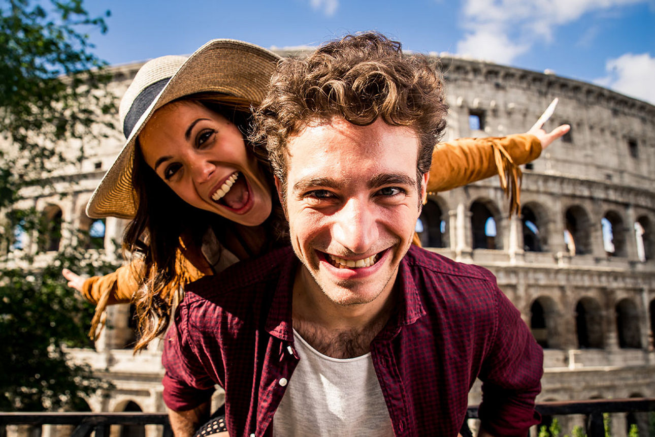 Eine Frau sitzt auf dem Rücken eines Mannes - im Hintergrund ist das Colloseum in Rom zu sehen.