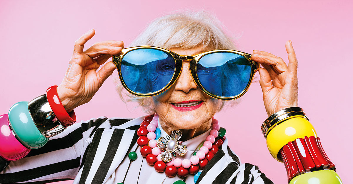 Eine ältere Dame zieht eine übergroße Brille auf. Die Dame trägt bunten, übergroßen Schmuck