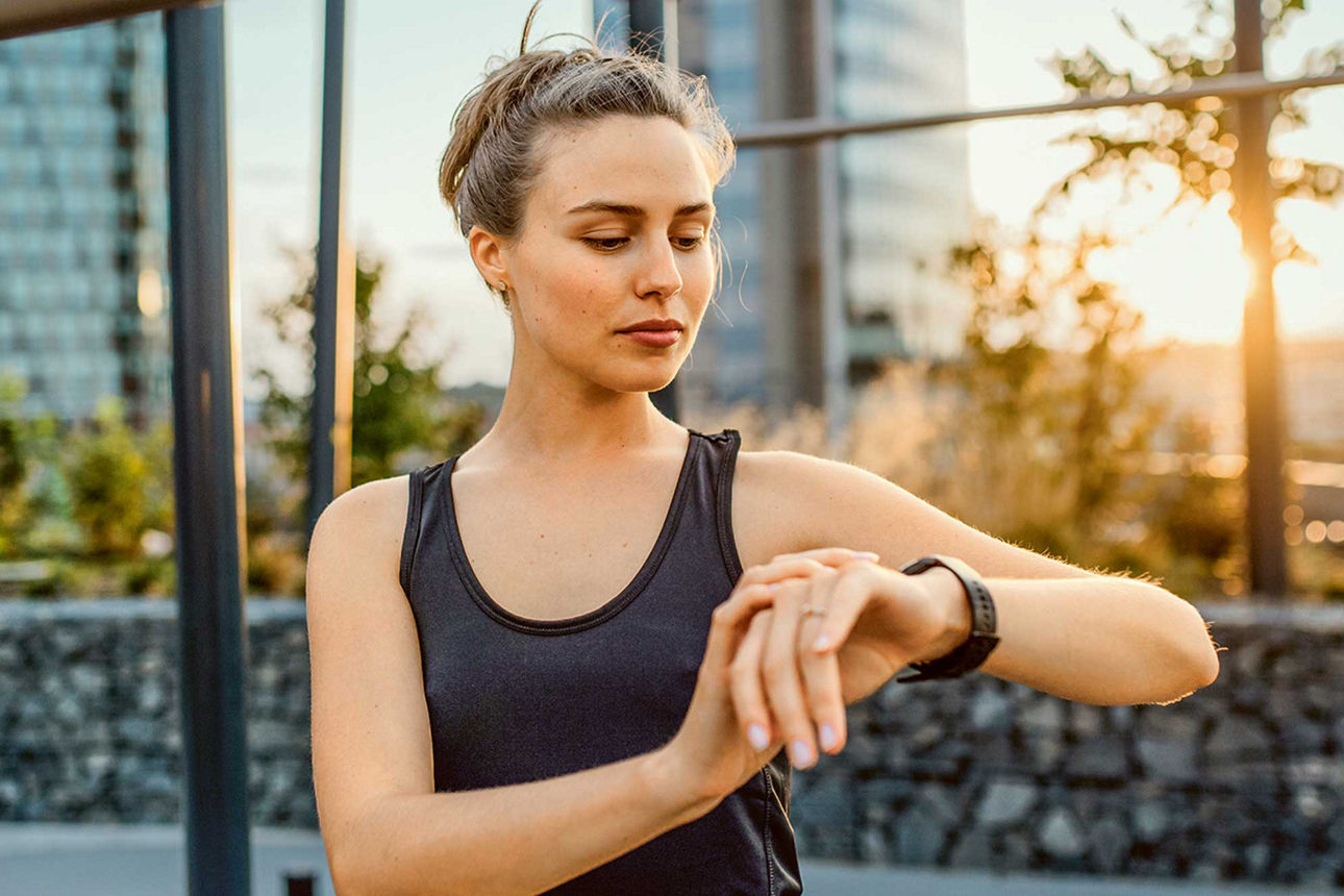 Eine Frau im Sportoutfit steht draußen und schaut auf ihre Smartwatch.