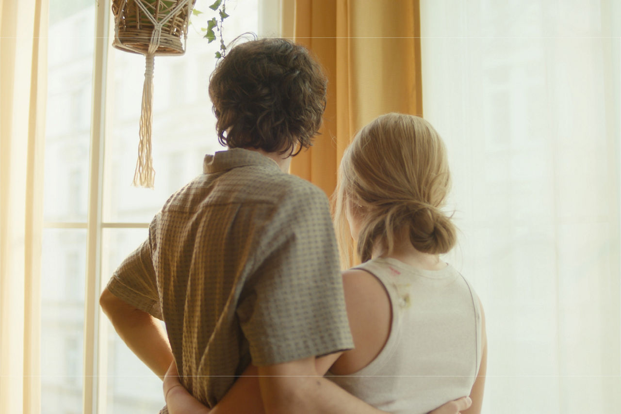Ein junges Pärchen steht Arm in Arm vor einem Fenster und blicken hinaus.