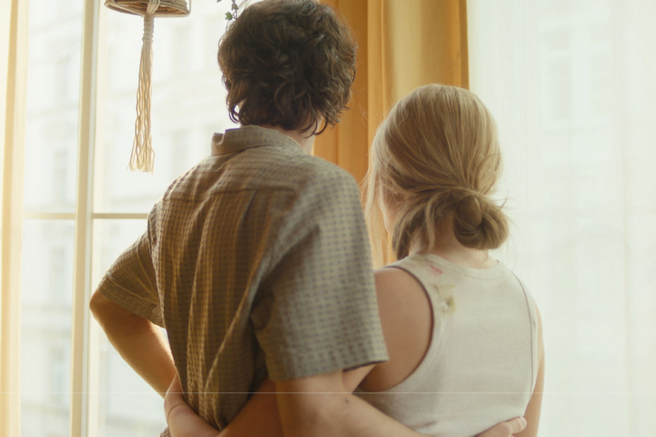 Ein junges Pärchen steht Arm in Arm vor einem Fenster und blicken hinaus.