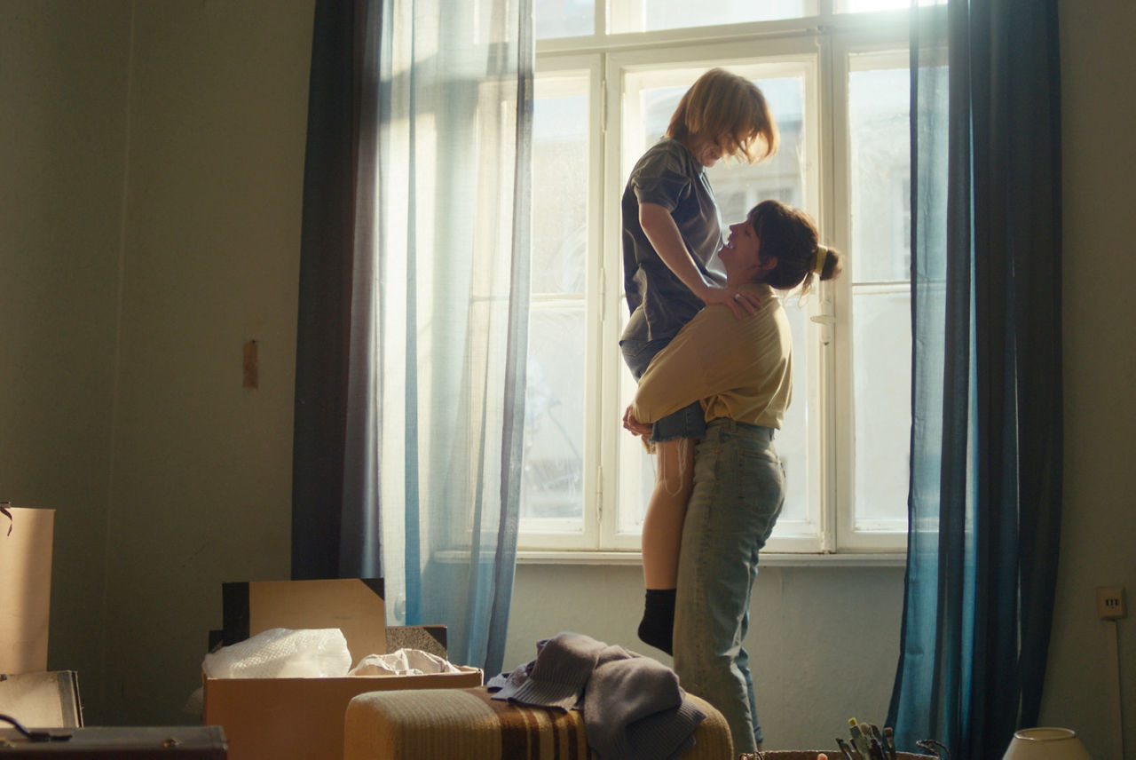 Zwei junge Frauen stehen in einer Wohnung mit Umzugskartons vor dem Fenster. Die eine hebt die andere voller Freude hoch.