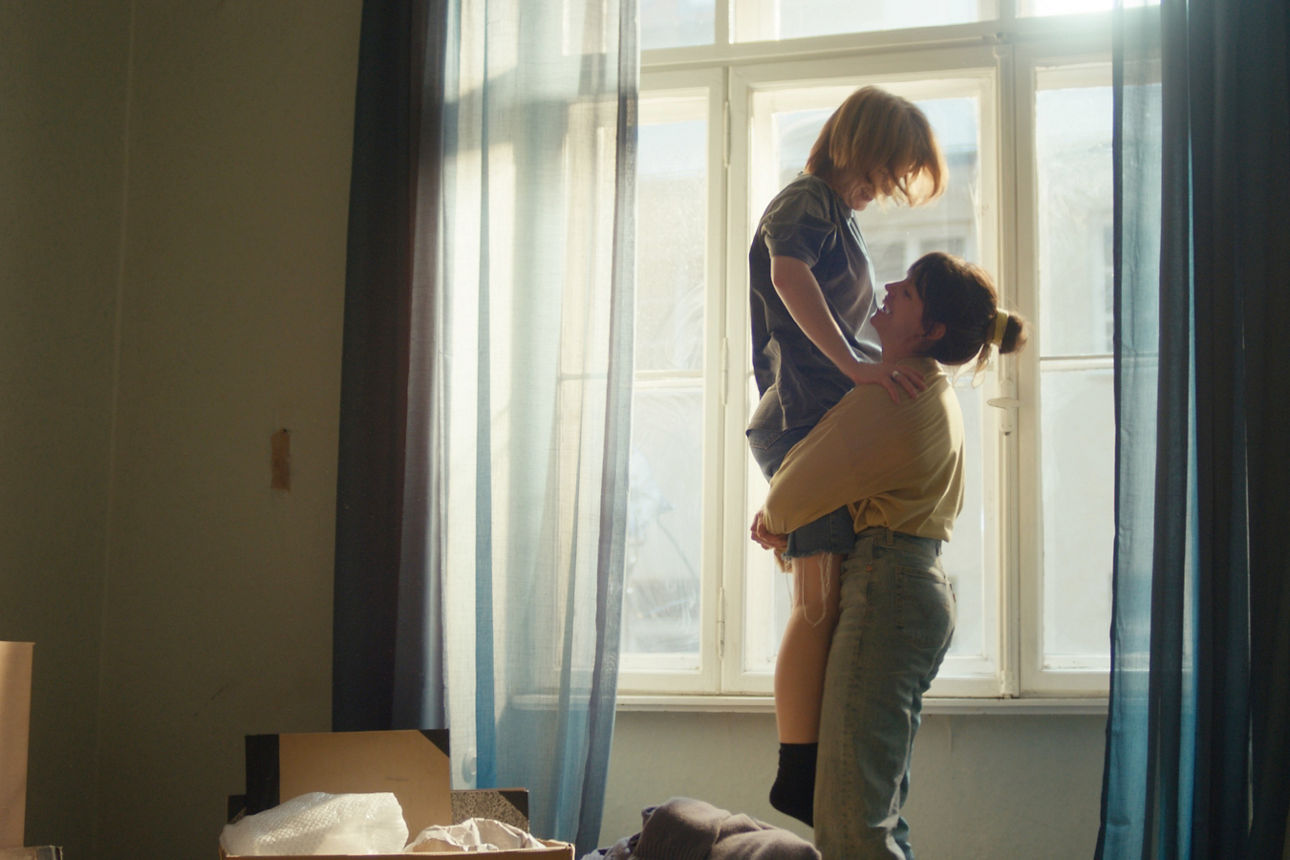 Zwei junge Frauen stehen in einer Wohnung mit Umzugskartons vor dem Fenster. Die eine hebt die andere voller Freude hoch.