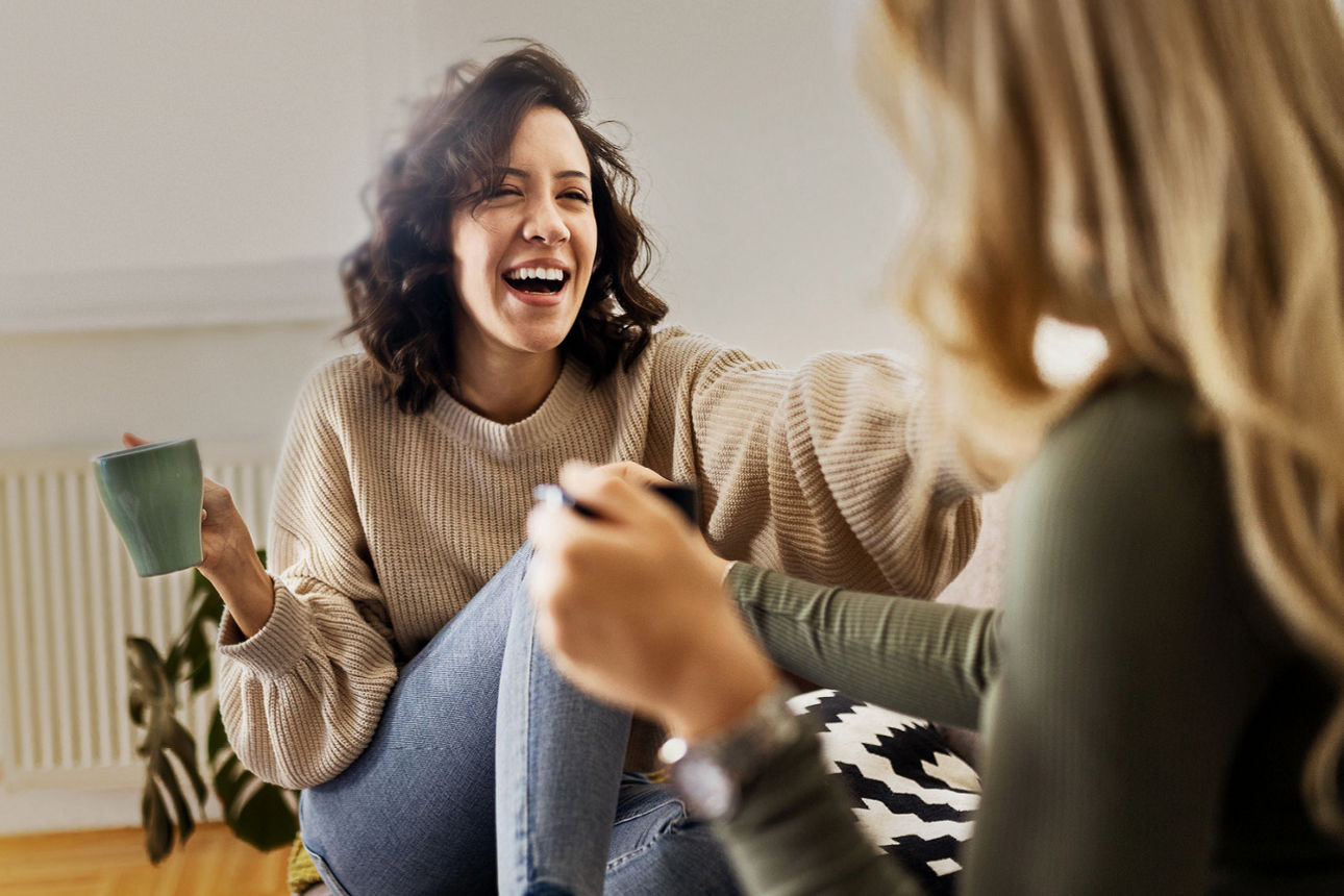 Zwei Frauen sitzen lachend mit einer Kaffeetasse in der Hand auf dem Boden