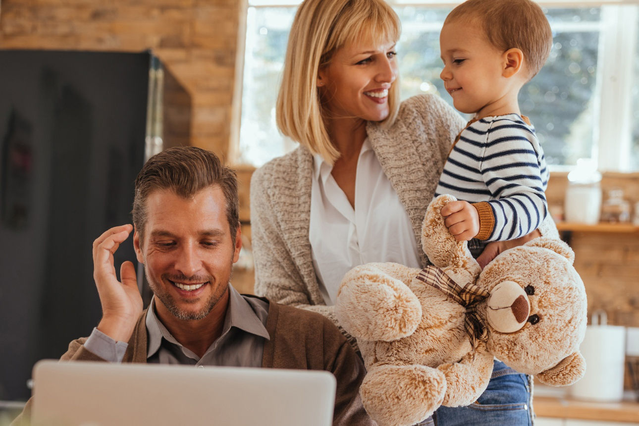 Junge Familie bestehend aus Vater, Mutter und Kleinkind. Vater sitzt am Notebook. Kleinkind hat Teedybär in der Hand.