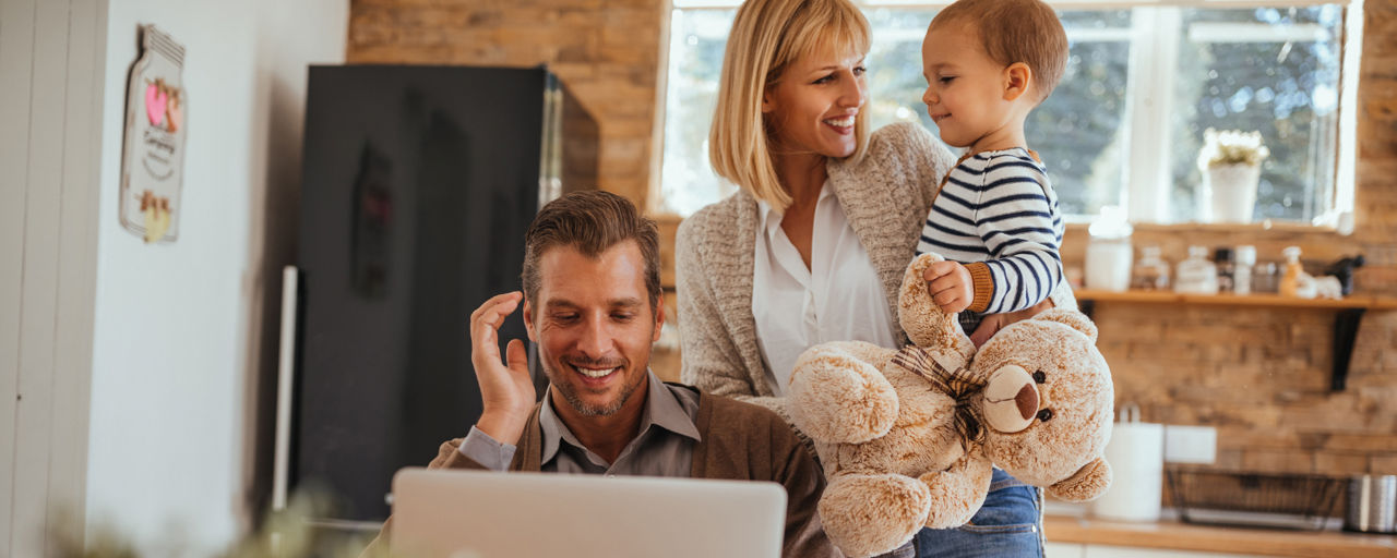 Junge Familie bestehend aus Vater, Mutter und Kleinkind. Vater sitzt am Notebook. Kleinkind hat Teedybär in der Hand.