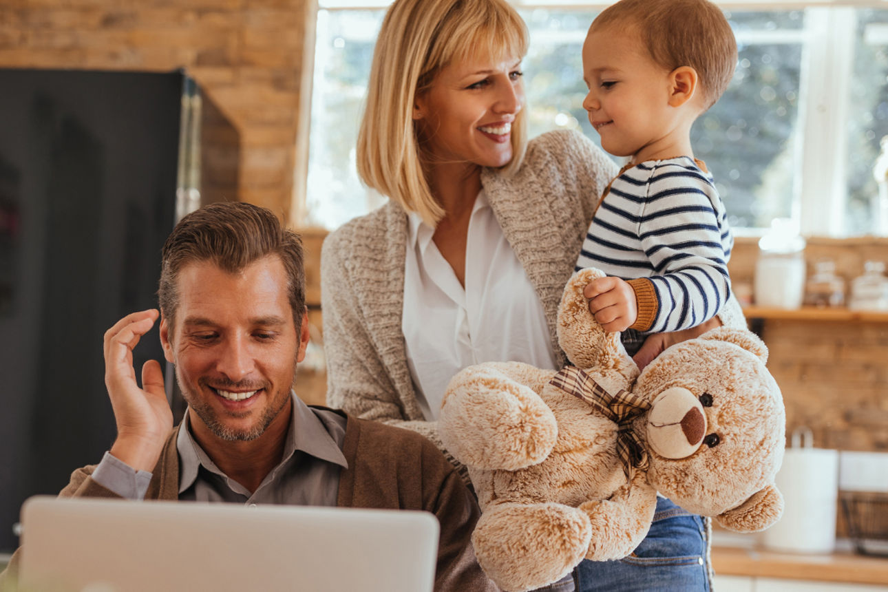 Junge Familie bestehend aus Vater, Mutter und Kleinkind. Vater sitzt am Notebook. Kleinkind hat Teedybär in der Hand.